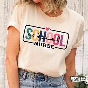 May include: A cream colored t-shirt with the text "SCHOOL NURSE" in a colorful block font with a small pink heart above the word "SCHOOL". The text is inside a black rectangle with a white outline. The shirt is being worn by a person.