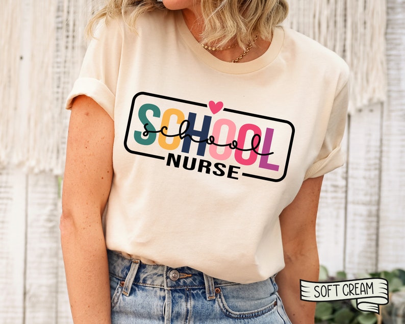 May include: A cream colored t-shirt with the text "SCHOOL NURSE" in a colorful block font with a small pink heart above the word "SCHOOL". The text is inside a black rectangle with a white outline. The shirt is being worn by a person.