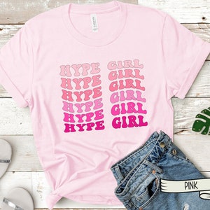 May include: A light pink t-shirt with the words "HYPE GIRL" in a retro font. The text is in shades of pink and magenta. The shirt is displayed on a wooden surface with accessories.