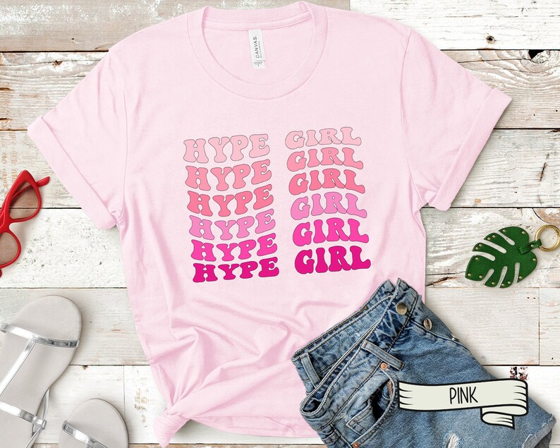 May include: A light pink t-shirt with the words "HYPE GIRL" in a retro font. The text is in shades of pink and magenta. The shirt is displayed on a wooden surface with accessories.