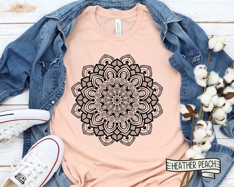May include: A peach-colored t-shirt with a black mandala design. The shirt is laid flat on a wooden surface with a denim jacket and cotton bolls.