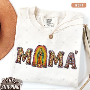 May include: Ivory t-shirt with a colorful graphic of the word "MOMA" with a depiction of the Virgin Mary in the center of the "O".