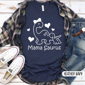 May include: A navy blue t-shirt with a white graphic of a mama dinosaur and a baby dinosaur with hearts. The text "Mama Saurus" is printed below the dinosaurs. The shirt is on a white wooden background with a plaid shirt, jeans, and white sneakers.