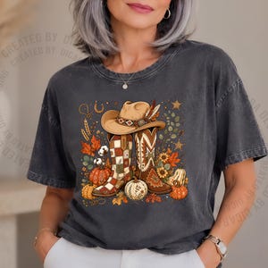 May include: A charcoal gray short-sleeve t-shirt featuring a graphic print of a tan cowboy hat resting on two western-style boots. The design includes autumn-themed elements such as orange pumpkins, colorful fall leaves, sunflowers, and stars.