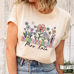 May include: A cream coloured t-shirt with a floral design and the text "Paraprofessional Mrs. Kate" in rainbow colours.