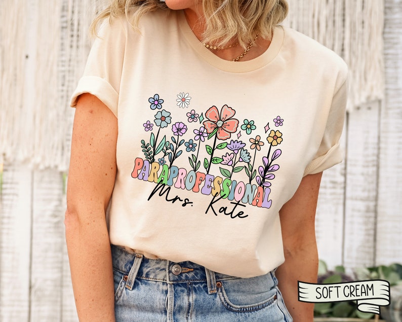 May include: A cream coloured t-shirt with a floral design and the text "Paraprofessional Mrs. Kate" in rainbow colours.