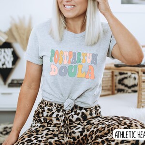 May include: A gray t-shirt with the text "POSTPARTUM DOULA" in colorful, retro-style lettering. The shirt is being worn by a person with long blonde hair and is sitting on a leopard print chair. The text "ATHLETIC HEATHER" is visible in the bottom right corner of the image.