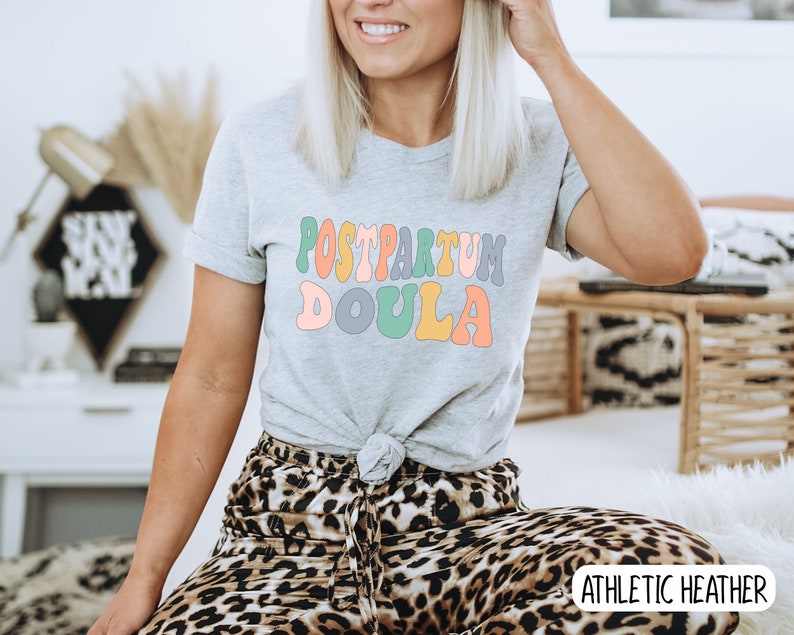 May include: A gray t-shirt with the text "POSTPARTUM DOULA" in colorful, retro-style lettering. The shirt is being worn by a person with long blonde hair and is sitting on a leopard print chair. The text "ATHLETIC HEATHER" is visible in the bottom right corner of the image.