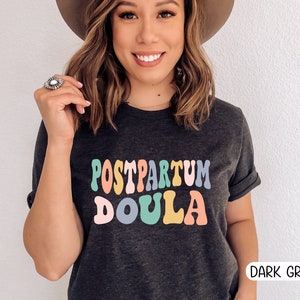 May include: A dark grey t-shirt with the text "POSTPARTUM DOULA" in colorful retro-style lettering.
