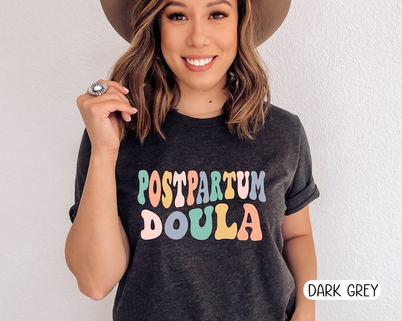 May include: A dark grey t-shirt with the text "POSTPARTUM DOULA" in colorful retro-style lettering.