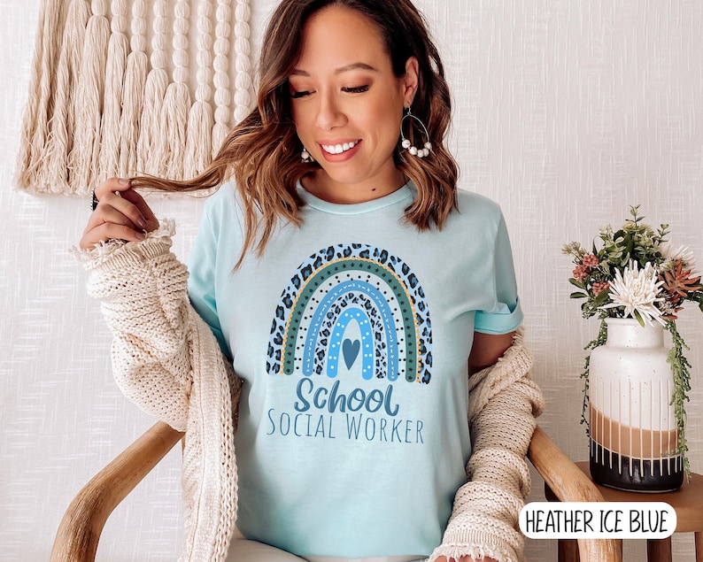 Puede incluir: Una camiseta azul claro con un dise&ntilde;o de arco&iacute;ris y el texto "School Social Worker". El arco&iacute;ris est&aacute; hecho de diferentes colores y tiene un coraz&oacute;n en el centro. La camiseta est&aacute; puesta en una modelo que lleva un c&aacute;rdigan.