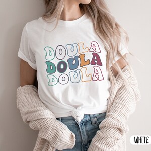 May include: White t-shirt with the word "Doula" repeated three times in a retro font with colorful outlines. The shirt is being worn by a person with long blonde hair.