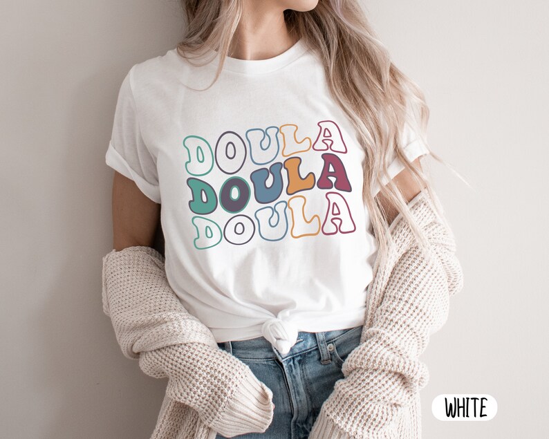 May include: White t-shirt with the word "Doula" repeated three times in a retro font with colorful outlines. The shirt is being worn by a person with long blonde hair.