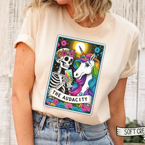 May include: A white t-shirt with a colorful illustration of a skeleton and a unicorn holding glasses. The illustration is in the style of a tarot card and says "The Audacity" at the bottom.