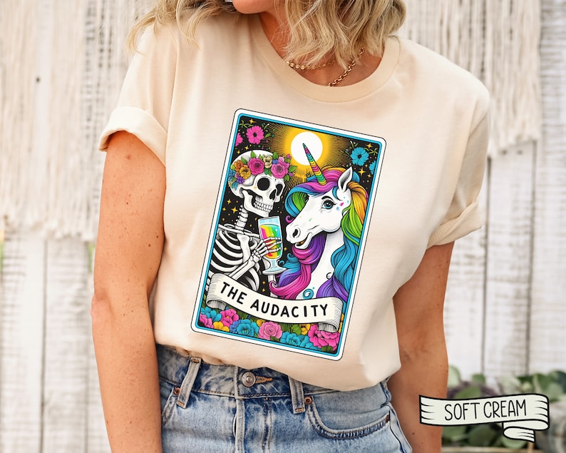 May include: A white t-shirt with a colorful illustration of a skeleton and a unicorn holding glasses. The illustration is in the style of a tarot card and says "The Audacity" at the bottom.