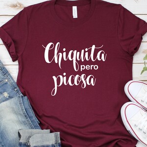 May include: A maroon t-shirt with the text "Chiquita pero picosa" in white cursive lettering.