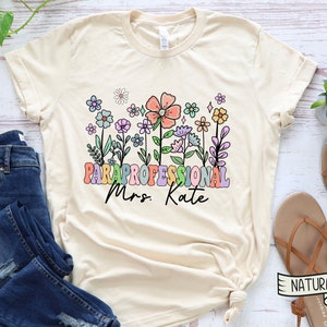 May include: A beige t-shirt with a floral design and the text "PARAPROFESSIONAL Mrs. Kate" in rainbow colours.