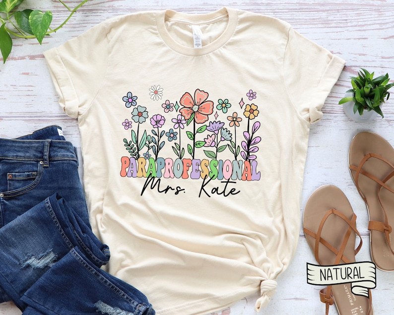May include: A beige t-shirt with a floral design and the text "PARAPROFESSIONAL Mrs. Kate" in rainbow colours.
