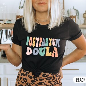 May include: Black t-shirt with the text "Postpartum Doula" in colorful, retro-style lettering. The shirt is worn by a person with blonde hair and is paired with a leopard print skirt.