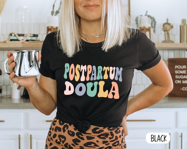 May include: Black t-shirt with the text "Postpartum Doula" in colorful, retro-style lettering. The shirt is worn by a person with blonde hair and is paired with a leopard print skirt.