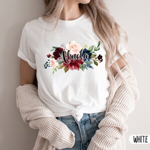 Pu&ograve; includere: Maglietta bianca con un design floreale e la parola "Abuela" in lettere nere.