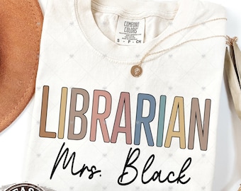 May include: Ivory-colored t-shirt with the word "LIBRARIAN" in colorful block letters and the name "Mrs. Black" in cursive. The shirt has a Comfort Colors tag and a gold necklace.