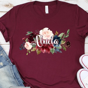 Pu&ograve; includere: Una maglietta color borgogna con un disegno floreale e la parola "Abuela" in lettere bianche.