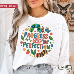 May include: White sweatshirt with a colorful graphic that reads "PROGRESS OVER PERFECTION". The design includes a caterpillar, butterflies, leaves, and a sun. The sweatshirt is a Comfort Colors brand.