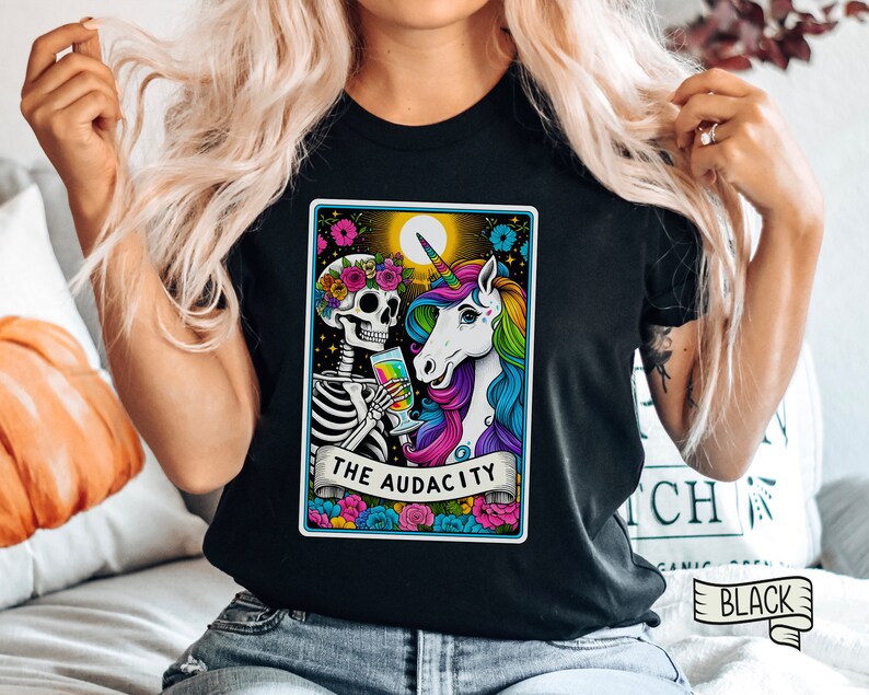 May include: Black t-shirt with a colorful illustration of a skeleton and a unicorn holding a glass of liquid. The illustration is framed in a card with the text "THE AUDACITY".