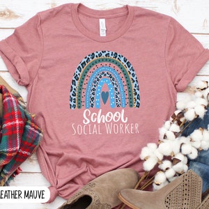 Puede incluir: Una camiseta de color malva claro con un dise&ntilde;o de arco&iacute;ris con estampado de leopardo azul, verde y marr&oacute;n. El arco&iacute;ris tiene un peque&ntilde;o coraz&oacute;n azul en el centro. El texto "School Social Worker" est&aacute; impreso debajo del arco&iacute;ris.