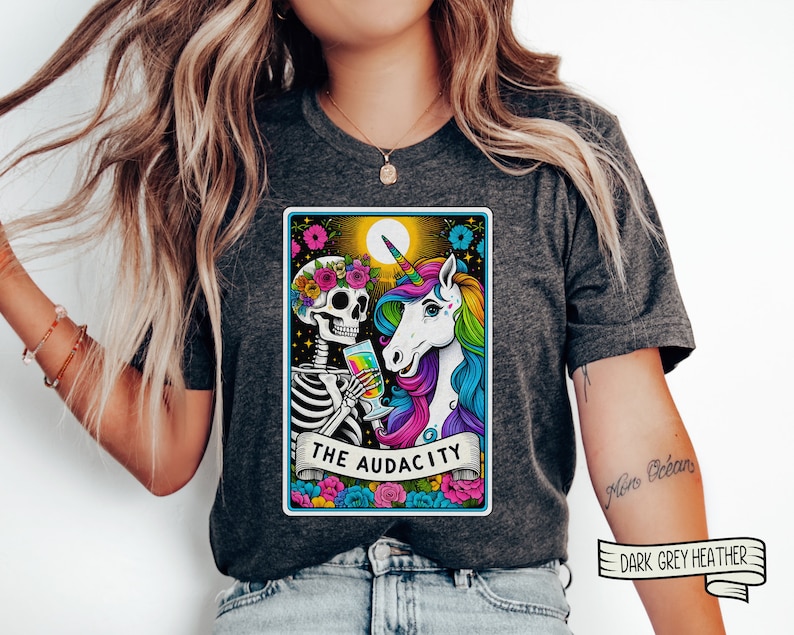 May include: A dark grey heather t-shirt with a colorful graphic of a skeleton and a unicorn holding glasses of liquid. The graphic is in a tarot card style with the text "The Audacity" at the bottom.