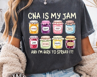 以下が含まれることがあります： 「CNA IS MY JAM」の文字と、「Kindness」、「Support」、「Love」などの言葉が書かれたカラフルなジャムの瓶のイラストが描かれた、濃いグレーのTシャツ。「AND I'M HERE TO SPREAD IT!」のフレーズが瓶の下に印刷されています。