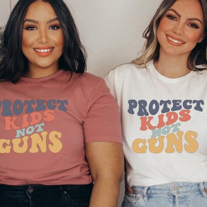 May include: Two people wearing t-shirts with the text "Protect Kids Not Guns" in a retro font with colorful letters. The shirts are pink and white.