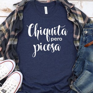 May include: A navy blue t-shirt with white text that reads "Chiquita pero picosa". The shirt is laid flat on a white wooden surface with a plaid shirt, a pair of jeans, and a pair of white sneakers.