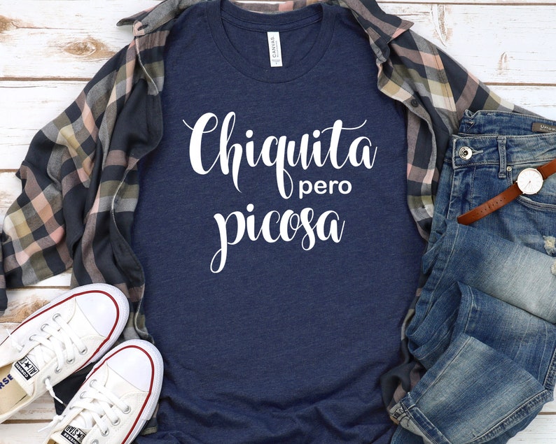 May include: A navy blue t-shirt with white text that reads "Chiquita pero picosa". The shirt is laid flat on a white wooden surface with a plaid shirt, a pair of jeans, and a pair of white sneakers.