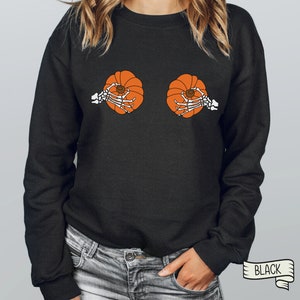 Op de afbeelding: Zwart sweatshirt met een afbeelding van twee skeletten die pompoenen vasthouden. De tekst "BLACK" is gedrukt op een wit lint.