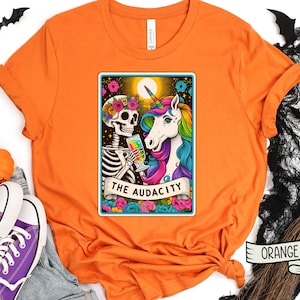 May include: Orange t-shirt with a graphic of a skeleton and a unicorn holding a glass of wine. The graphic is in a tarot card style with the text "The Audacity".