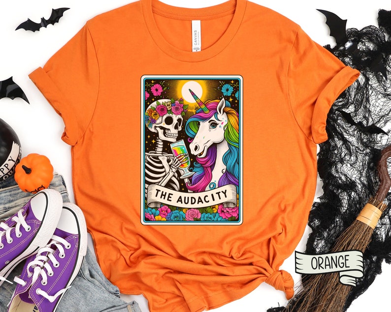 May include: Orange t-shirt with a graphic of a skeleton and a unicorn holding a glass of wine. The graphic is in a tarot card style with the text "The Audacity".
