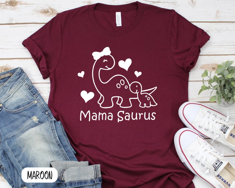 May include: Maroon t-shirt with a white outline of a mama dinosaur and baby dinosaur with hearts. The text "Mama Saurus" is printed below the dinosaurs.