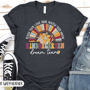 May include: Dark grey heather t-shirt with a rainbow design and the text "TEACH THEM, LOVE THEM, WATCH THEM GROW KINDERGARTEN dream team".