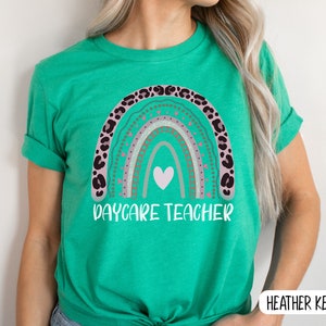 May include: A green t-shirt featuring a rainbow design and the text "Daycare Teacher". The rainbow is decorated with pink, brown, and white accents.
