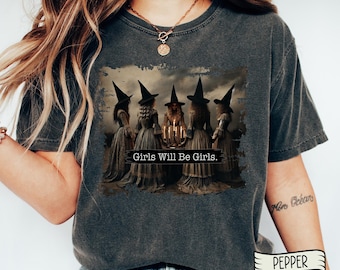 May include: A dark gray t-shirt with a vintage-style graphic of four witches standing around a cauldron with candles. The text "Girls Will Be Girls." is printed below the graphic.