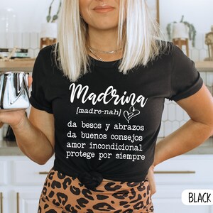 May include: Black t-shirt with white text that reads "Madrina [madre-nah] da besos y abrazos da buenos consejos amor incondicional protege por siempre".
