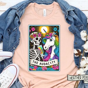 May include: A peach-colored t-shirt with a graphic of a skeleton and a unicorn holding drinks. The graphic is in a tarot card style with the text "THE AUDACITY" at the bottom. The t-shirt is on a wooden surface with a denim jacket and cotton bolls.