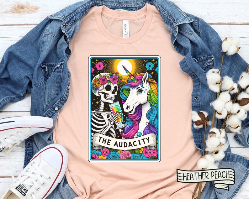 May include: A peach-colored t-shirt with a graphic of a skeleton and a unicorn holding drinks. The graphic is in a tarot card style with the text "THE AUDACITY" at the bottom. The t-shirt is on a wooden surface with a denim jacket and cotton bolls.