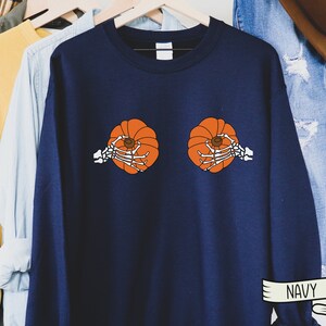 Op de afbeelding: Een marineblauw sweatshirt met een afbeelding van twee pompoenen die door skelethandjes worden vastgehouden. De tekst "NAVY" is afgedrukt op een wit lint aan de onderkant van de afbeelding.