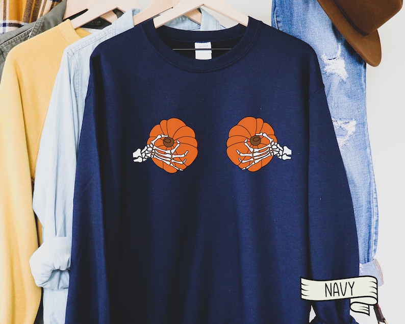 Op de afbeelding: Een marineblauw sweatshirt met een afbeelding van twee pompoenen die door skelethandjes worden vastgehouden. De tekst "NAVY" is afgedrukt op een wit lint aan de onderkant van de afbeelding.