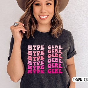 May include: Dark grey t-shirt with the text "HYPE GIRL" in a pink and fuchsia retro font. The shirt has rolled-up sleeves and is a casual style. The words "DARK GREY" are printed in a white font at the bottom right.