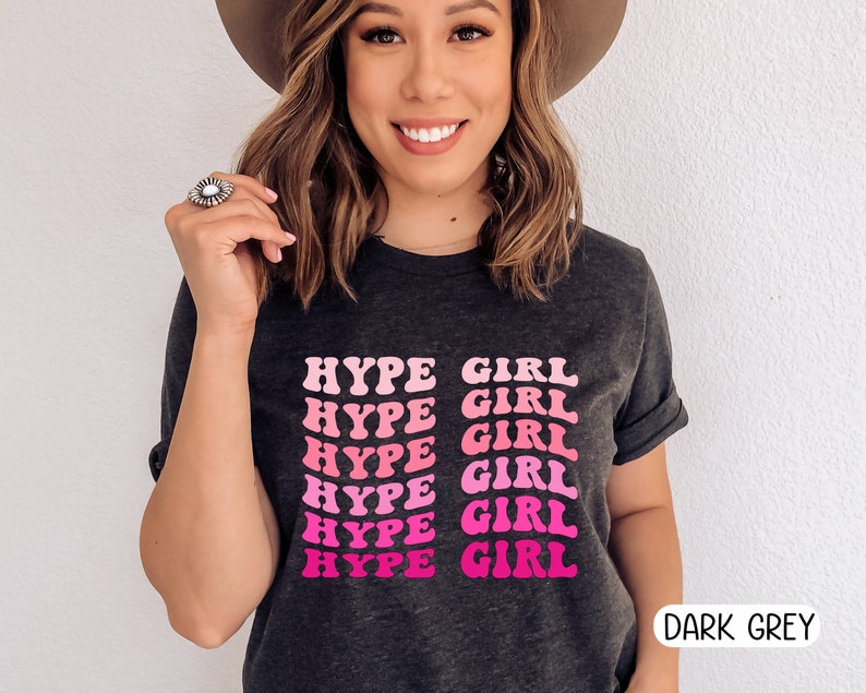 May include: Dark grey t-shirt with the text "HYPE GIRL" in a pink and fuchsia retro font. The shirt has rolled-up sleeves and is a casual style. The words "DARK GREY" are printed in a white font at the bottom right.