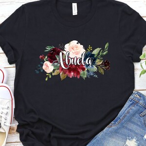 Pu&ograve; includere: Maglietta nera con un design floreale e la parola "Abuela" in corsivo bianco.